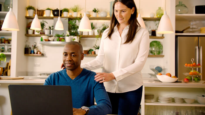 Foto på en man som sitter ned i köket och hans fru står upp bredvid honom. Båda tittar på en bärbar dator