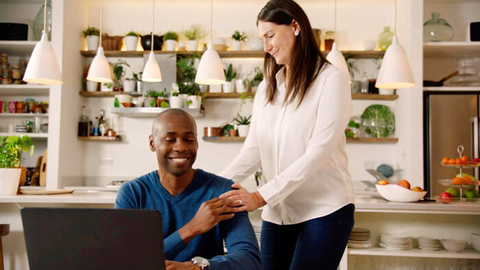 Foto på en man som sitter ned i köket och ler och hans fru står upp bredvid honom och ler mot honom