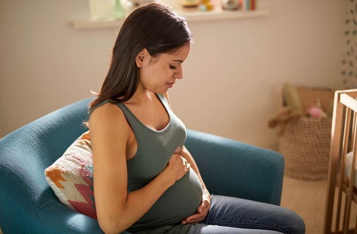 En gravid kvinna sitter i soffan med slutna ögon och håller sig om magen.