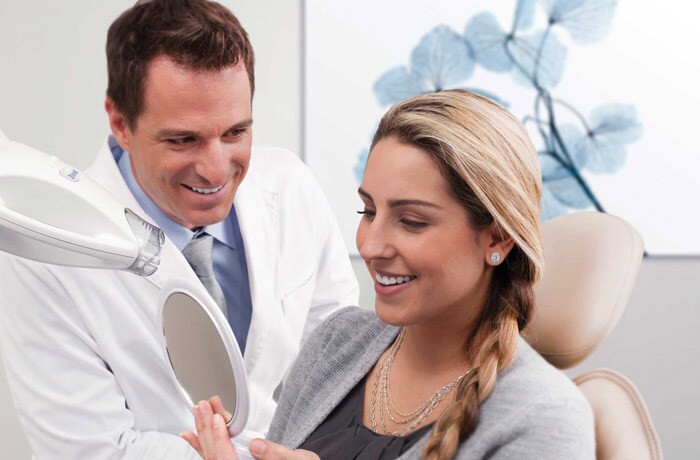 En kvinna ler och visar sina vita tänder i spegeln samtidigt som hon pratar med sin tandhygienist.