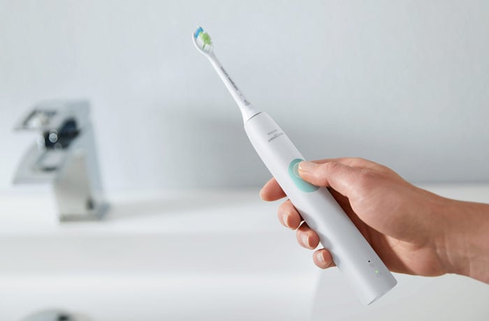 En hand som håller en vit Philips Sonicare eltandborste framför ett handfat med tummen på knappen.