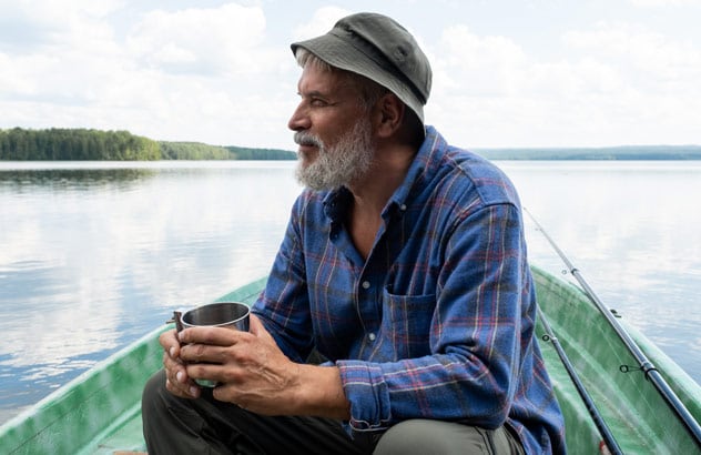 En äldre man med grått skägg sitter i en båt ute på vattnet iklädd en blå skjorta och grön hatt, hållandes en kopp. 
