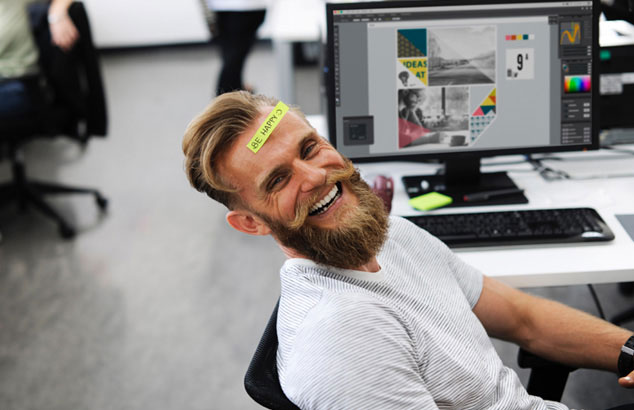En man med hipsterskägg och en post-it med orden "Be happy" i pannan som sitter på ett kontor.