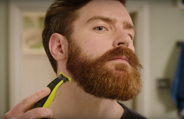 En man trimmar ner sitt rufsiga skägg till en skäggstubb med hjälp av en trimmer med specialtillbehör.