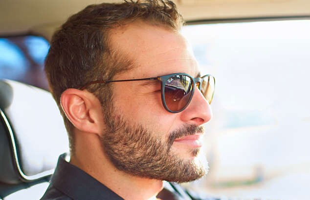 Sidoprofil av en man som sitter i en bil med kort helskägg och solglasögon.