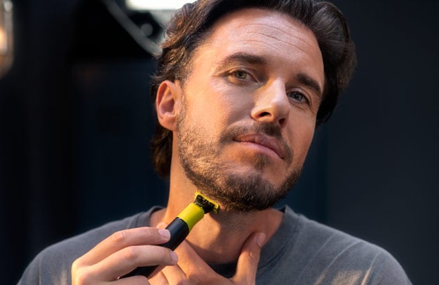 Närbild av en man som trimmar kanterna på sitt mörka skägg med Philips OneBlade framför en spegel.