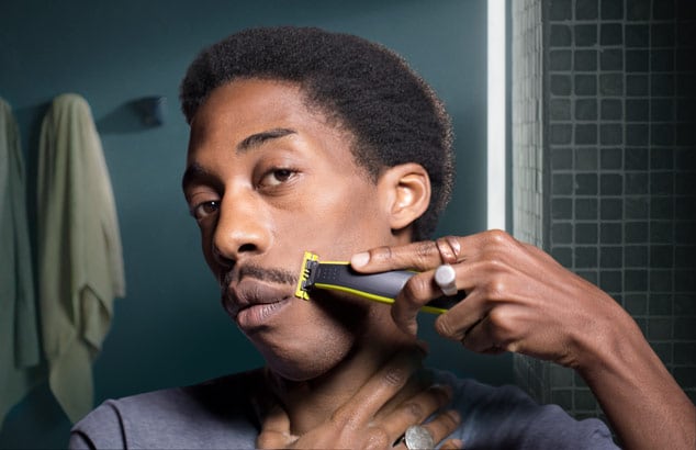 En ung mörkhyad man trimmar mustaschen framför badrumsspegeln med Philips OneBlade.