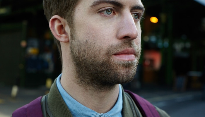 Närbild av en man med tredagarsskägg som tittar in i kameran från sidan.